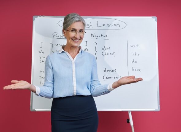 Cheerful senior woman teaching English language near the whiteboard against pink background